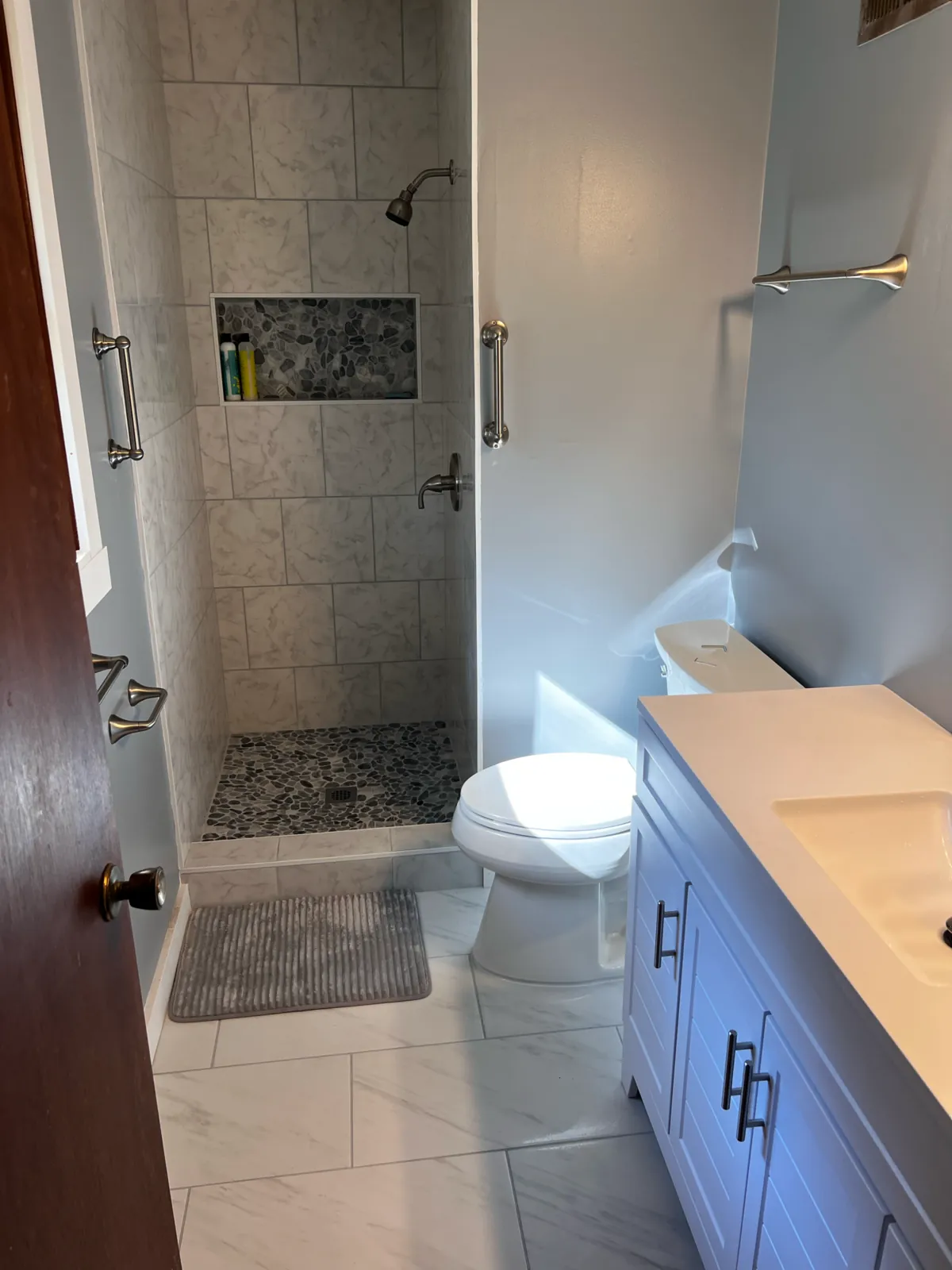 Marble-look tile walk-in shower with a stone niche, pebble accent, grab bars, and a vanity alongside